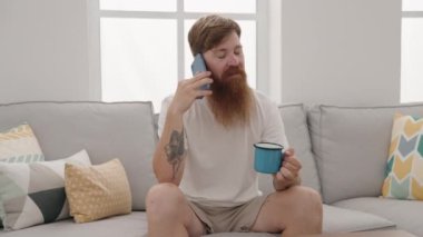 Young redhead man talking on the smartphone sitting on sofa at home