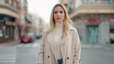 Genç sarışın kadın gülümsüyor. Kollarını kavuşturup caddede duruyor.