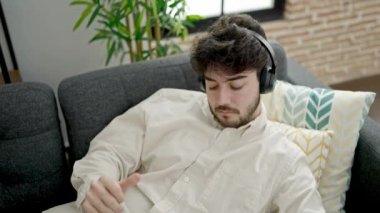 Genç İspanyol adam evde kanepede uzanmış akıllı telefonuyla müzik dinliyor.
