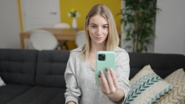 Genç sarışın kadın telefonuyla video görüşmesi yapıyor. Evdeki kanepede oturuyor.