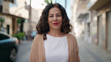 Orta yaşlı kadın gülümsüyor. Kollarını kavuşturup caddede duruyor.