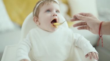 Sevimli sarışın bebek sandalyeye oturmuş, evde yemek yiyor.