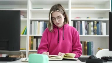 Young blonde woman student reading book doing silence gesture at library university