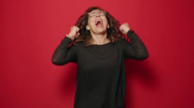 Orta yaşlı kadın gülümsüyor. Kırmızı arka planda kutlama yaparken kendinden emin duruyor.