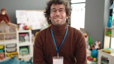 Young hispanic man preschool teacher smiling confident standing with arms crossed gesture at kindergarten