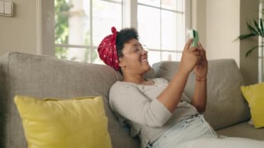 Evdeki koltukta oturan Afro-Amerikan kadın akıllı telefon kullanıyor