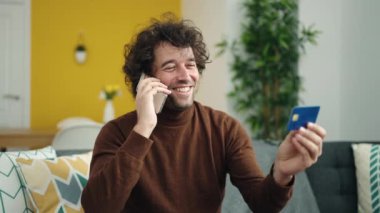 Young hispanic man talking on smartphone and credit card sitting on sofa at home