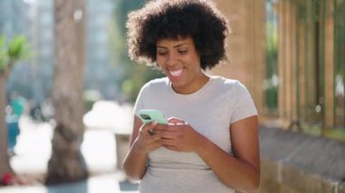 Genç Afro-Amerikalı kadın sokakta akıllı telefon kullanarak gülümsüyor.