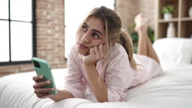 Young blonde woman using smartphone with sad expression at bedroom