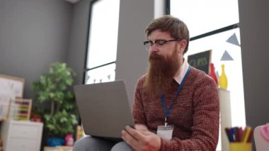 Young redhead man preschool teacher using laptop sitting on chair at kindergarten