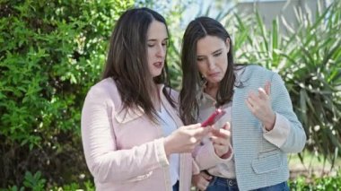 Kendinden emin gülümseyen iki kadın parkta akıllı telefondan selfie çekiyor.