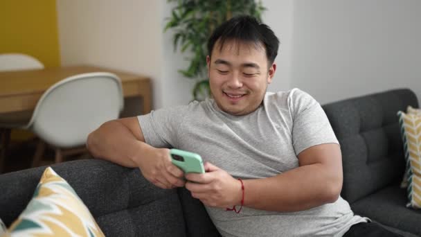 Young Chinese Man Using Smartphone Sitting Sofa Home — стокове відео