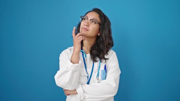 若いです美しいですヒスパニック女性医師立っています疑い式考えます上の隔離された青い壁の背景 — ストック動画