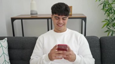 Young hispanic man using smartphone doing ok gesture at home