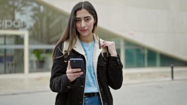 Young beautiful hispanic woman student smiling confident using smartphone at street