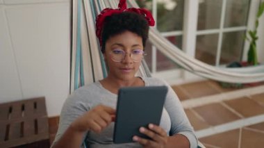 Ev terasında hamakta uzanmış dokunmatik ped kullanan Afrikalı Amerikalı bir kadın.