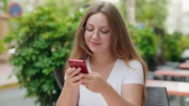 Genç sarışın kadın kahve terasında masada oturan akıllı telefonu kullanıyor.