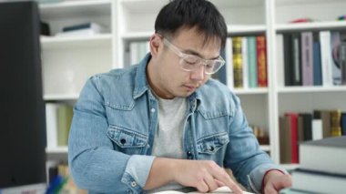 Young chinese man student reading book sitting on table at library university