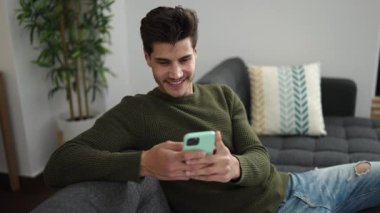 Young hispanic man using smartphone doing ok gesture at home