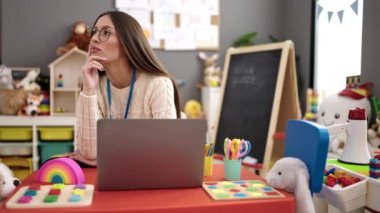 Young beautiful hispanic woman preschool teacher using laptop sitting on table at kindergarten