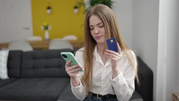 年轻的金发女人坐在家里的沙发上 手里拿着智能手机和信用卡购物 — 图库视频影像