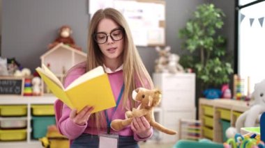 Young blonde woman preschool teacher reading story book at kindergarten