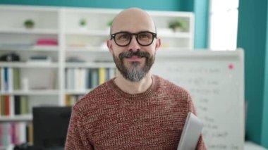 Genç ve kel bir öğretmen üniversite sınıfında kendini kameraya göstererek gülümsüyor.