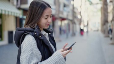 Genç, güzel, İspanyol bir kadın sokakta akıllı telefon kullanarak gülümsüyor.