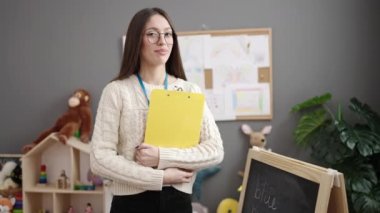 Young beautiful hispanic woman preschool teacher smiling confident holding clipboard at kindergarten