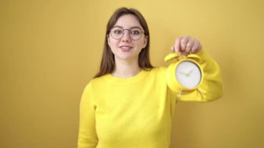 Young blonde woman smiling confident pointing with finger to alarm clock over isolated yellow background