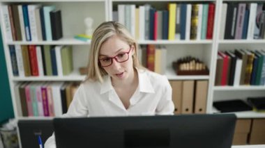 Young blonde woman student using computer writing on notebook at library university