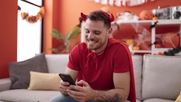 Young Caucasian Man Wearing Devil Costume Sitting Sofa Using Smartphone — Stockvideo