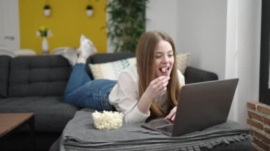 Young blonde woman watching movie lying on sofa at home
