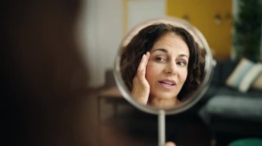 Middle age hispanic woman sitting on sofa looking face on mirror at home