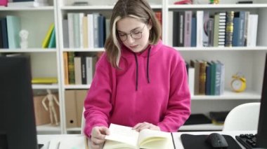 Young blonde woman student reading book sitting on table at library university