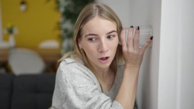 Young blonde woman listening through wall with glass at home