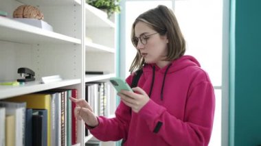 Kütüphanede akıllı telefon kullanarak kitap arayan genç sarışın kız öğrenci.