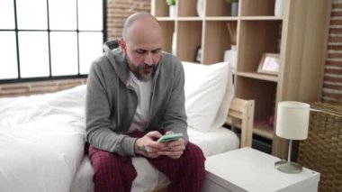 Genç kel adam yatakta akıllı telefon kullanıyor.