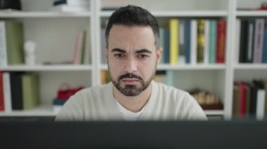 Young hispanic man student using computer with doubt expression at library university