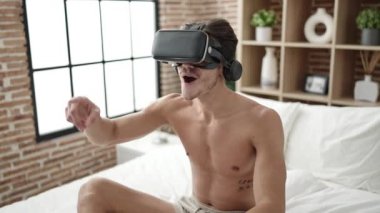 Young hispanic man sitting on bed using virtual reality glasses at bedroom