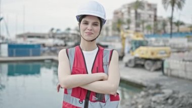 Genç, güzel İspanyol kadın müteahhit, gülümsüyor. Sahilde kollarını kavuşturmuş duruyor.