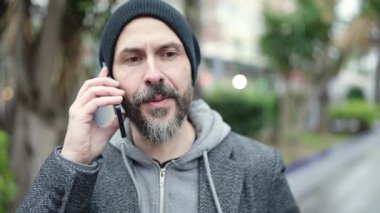 Genç kel adam gülümsüyor. Parkta akıllı telefondan konuşuyor.