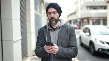 Genç kel adam akıllı telefon kullanıyor ve sokakta ciddi bir yüz ifadesi var.