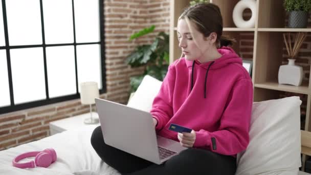 年轻的金发女人坐在卧室的床上 用笔记本电脑和信用卡 — 图库视频影像