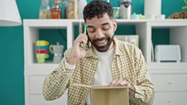 Genç İspanyol adam, yemek odasındaki karton kutuları açarak akıllı telefondan konuşuyor.