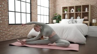Young blonde woman sitting on floor stretching legs at bedroom