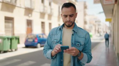 Young hispanic man using smartphone with winner gesture at street