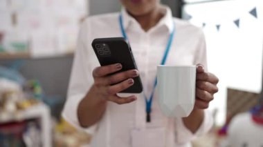 African american woman preschool teacher using smartphone drinking coffee at kindergarten