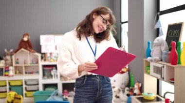 Young caucasian woman working as teacher writing on clipboard speaking on the telephone at kindergarten