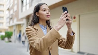 Genç, güzel, İspanyol bir kadın akıllı telefonuyla sokakta gülümseyen bir video kaydediyor.